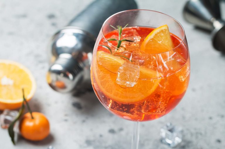 Ein alkoholfreier Aperol Spritz mit einer attraktiver, leuchtend orange Farbe in einem großen, bauchigen Weinglas mit Eiswürfeln. Eine Scheibe Orange und ein Rosmarinzweig als Garnierung. Im Hintergrund typisches Cocktailzubehör.