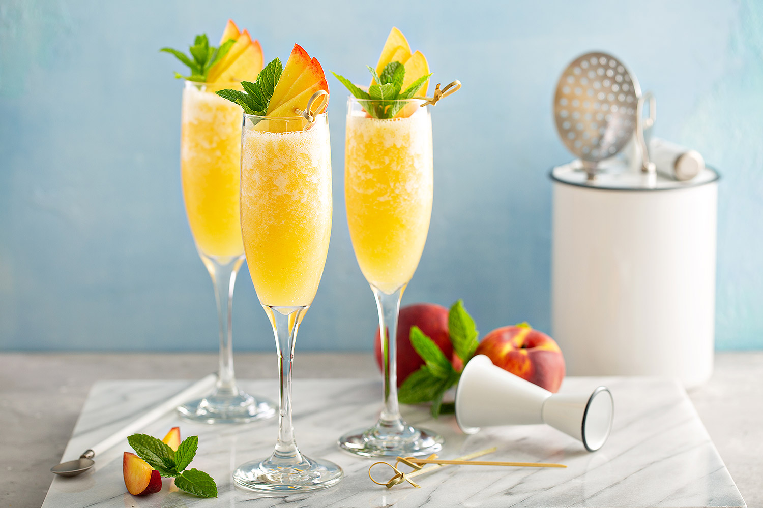 Three Bellini cocktails in champagne glasses on a white marble platter and garnished with mint leaves and peach slices.