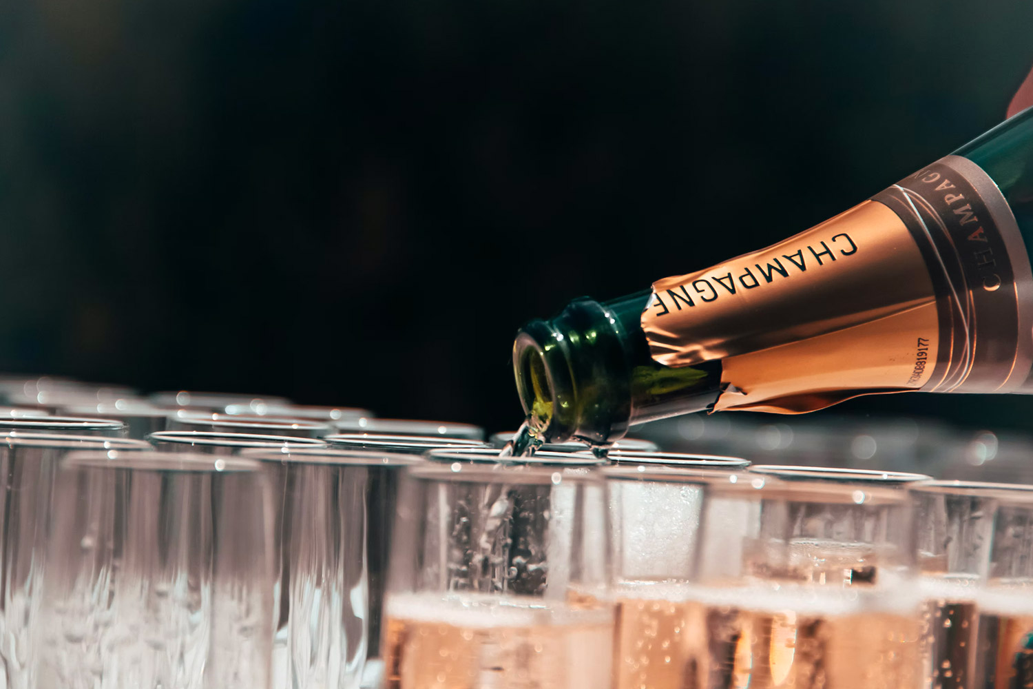 A bottle of champagne is poured on several glasses standing in front of a black background