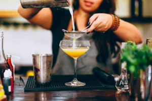 Bartender seiht einen gelben Cocktail mit 3 Zutaten in ein Glas ab.