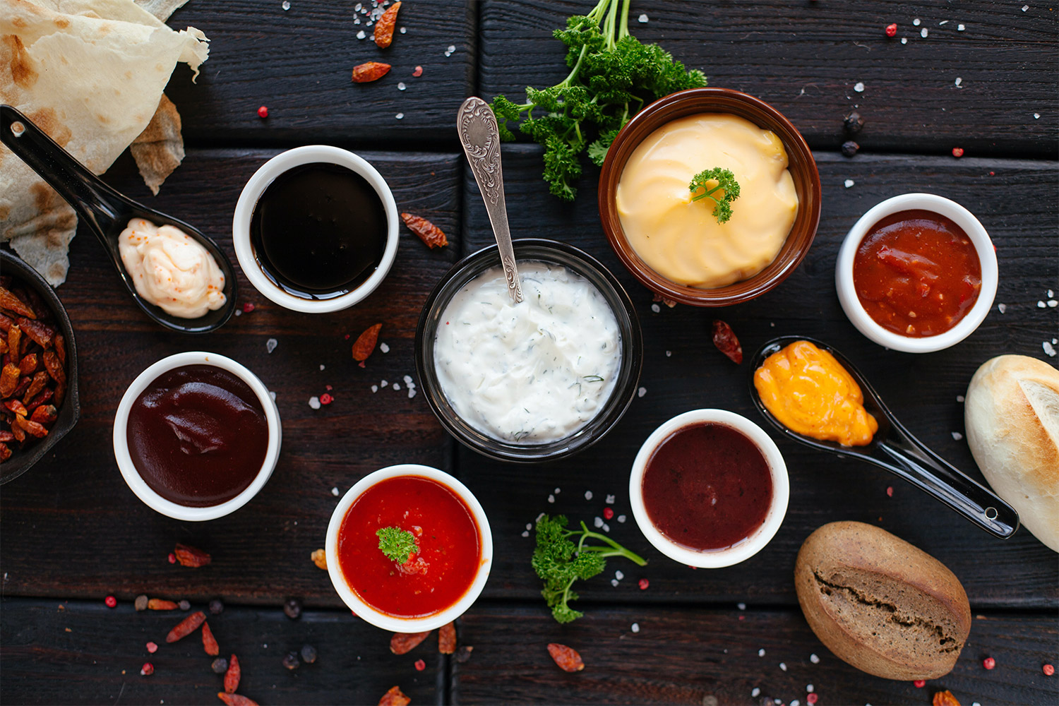 Seven different dips on a dark wooden board with spices and herbs scattered around it