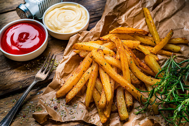 Selbst gemachte Pommes frites auf Backpapier mit Ketchup, Mayonnaise und Kräutern auf einem kleinen Holzbrett