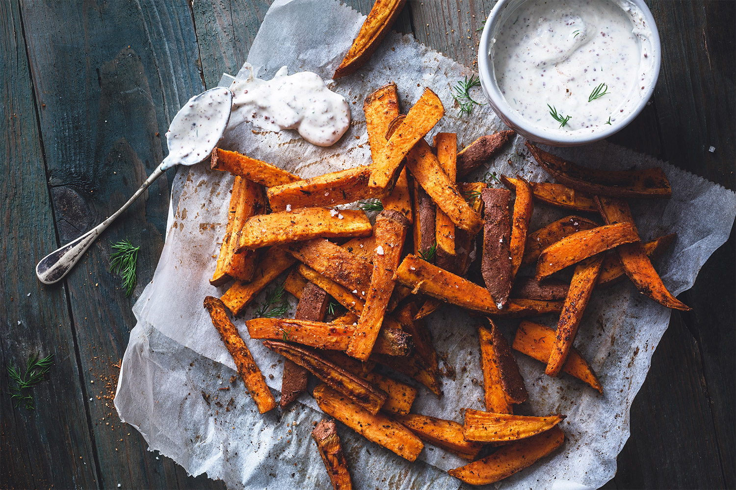 Sweet potato fries on white baking paper, with garlic dip on dark wooden boards.