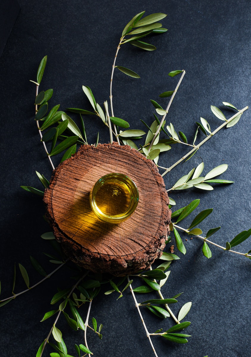 Olivenöl in einem kleinen Glas, auf einem Holzbrett vor dunklem Hintergrund mit Olivenzweigen als Deko.