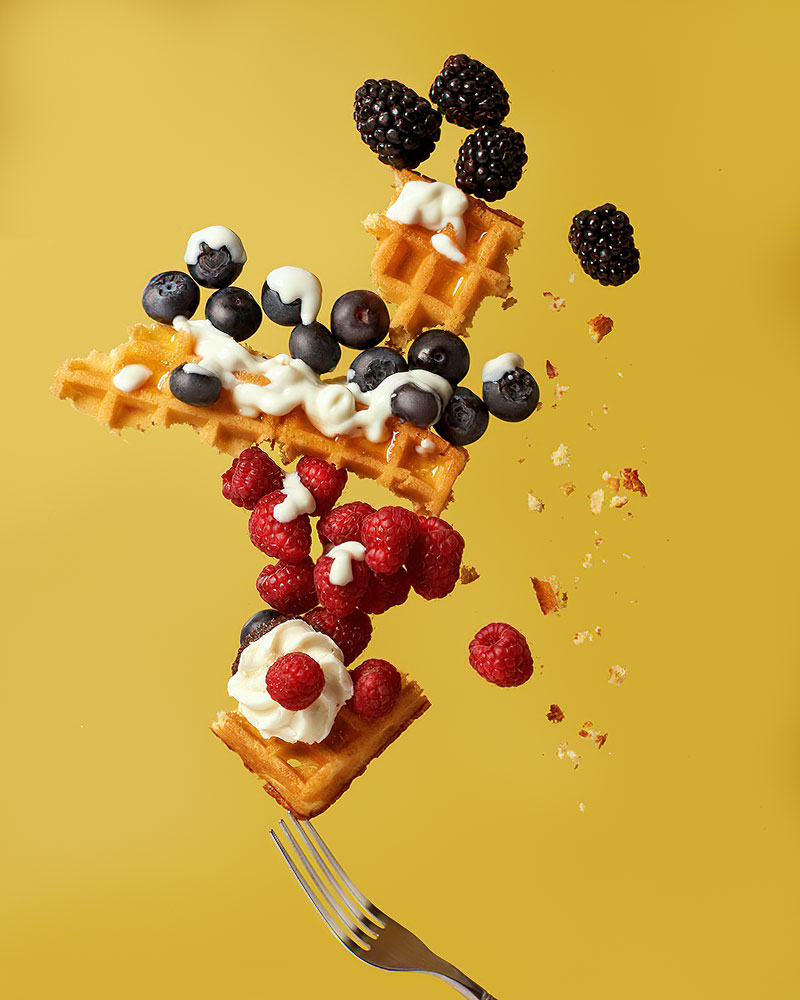 A broken wafer with cream, raspberries, blackberries and blueberries flies through the air against a yellow background.