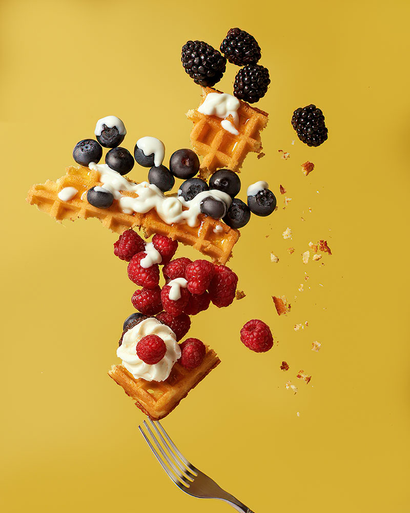 Eine zebrochene Waffel mit Sahne, Himbeeren, Brombeeren und Heidelbeeren fliegt vor einem gelben Hintergrund durch die Luft.