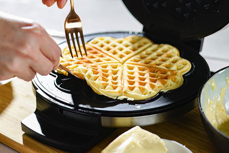 A finished waffle, baked golden brown, is lifted out of a hot waffle iron with a fork.