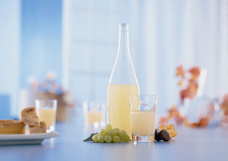 Eine Flasche und ein Glas Federweißer vor hellem Hintergrund und symbolischen Weintrauben im Vordergrund
