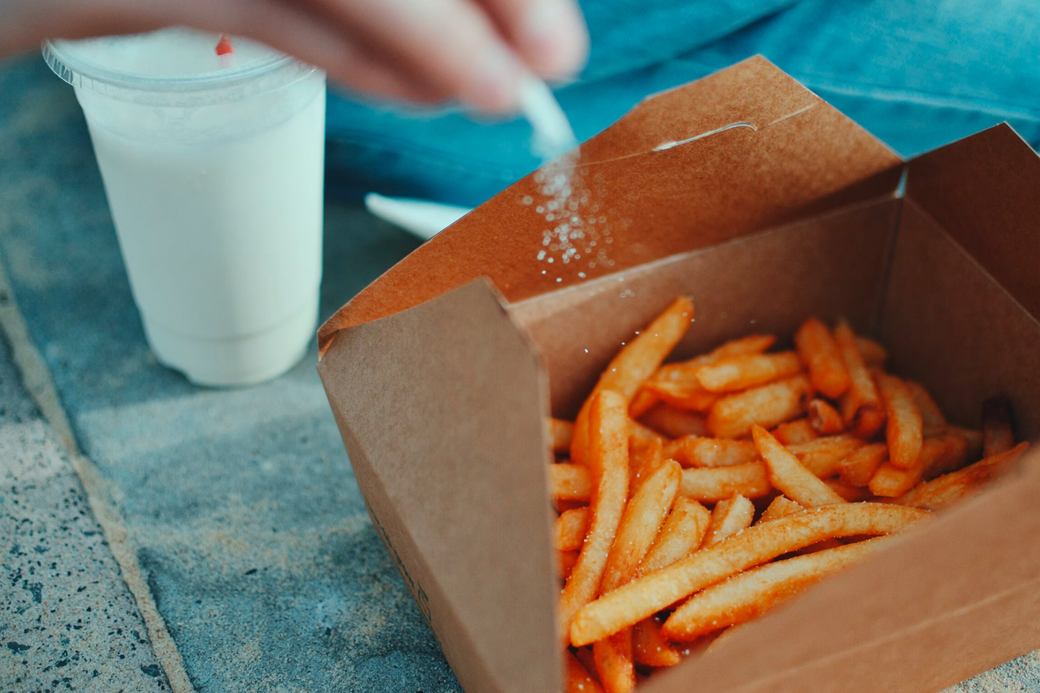 French fries served in a cardboard box on blue tiles and a hand sprinkling salt over them